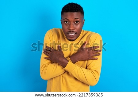 Desperate young handsome man wearing yellow sweater over blue background trembles and feels cold, hugs oneself to warm up or feels scared notices something terrifying.