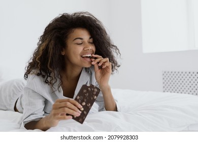 Dessert Food And Guilty Pleasure Concept. Happy Woman Eating Chocolate With Nuts While She Lying In Bed, Wearing In Pajamas. Smiling Female Biting Candy Bar, Enjoying Taste, Spending Morning At Home