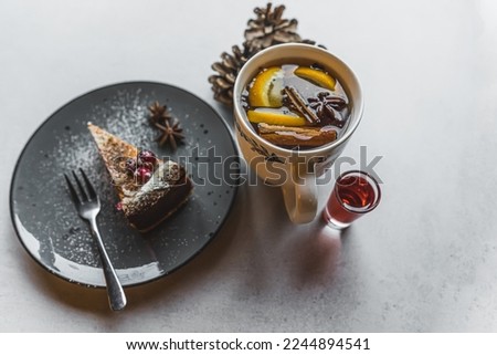 Dessert and a hot drink. A piece of cake decorated with powdered sugar and fruits placed on a black plate next to a white cup with a hot wintry drink and a small glass of red syrup. High quality photo