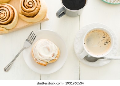 Dessert Is Served. Flat Lay Of A Glazed Cinnamon Roll On A White Plate Next To A Hot Cup Of Coffee Placed On A Wooden Kitchen Table 