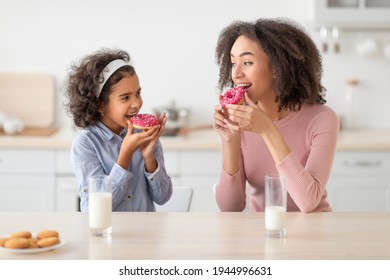 Dessert Time For Sweet Tooth. Portrait Of Positive Black Little Girl And Young Woman Eating Delicious Donuts Cakes And Looking At Each Other, Sitting At Dinner Table, Enjoying Breakfast With Milk