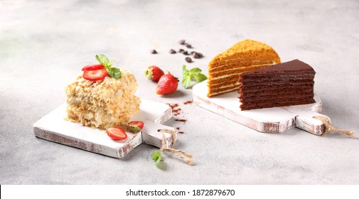 Desserts. Cakes: Napoleon, honey cake and chocolate cake on light wooden boards on a light gray table. Fresh berries, strawberries and mint, spoon. Background image, copy space, horizontal