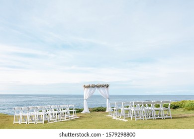 Destination  Beach Wedding Ceremony Decor - White Square Arch With Floral Arrangements, White Chairs  On The Lawn And Pacific Beach As A Background