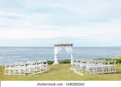 Destination  Beach Wedding Ceremony Decor - White Square Arch With Floral Arrangements, White Chairs  On The Lawn And Pacific Beach As A Background