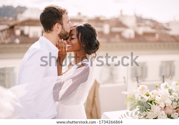 Destination fine-art wedding\
in Florence, Italy. Caucasian groom and African-American bride\
cuddling on a rooftop in sunset sunlight. Multiracial wedding\
couple