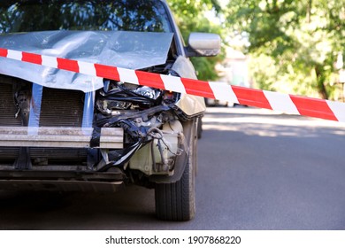 Destroyed Car In Car Crash Traffic Accident On City Road With Red Warning Police Tape. Smashed Broken Front Auto Headlight, Dented Hood Without Bumper On Gray Car Accident.