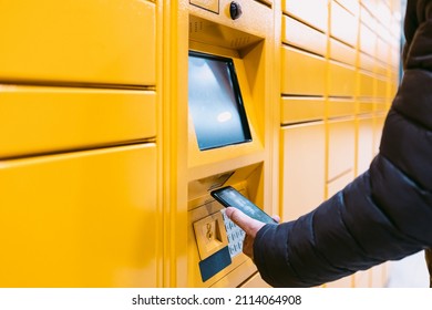 Detail Of The Arm Of A Man Scanning A Code On The Mobile Phone To Pick Up A Package From The Yellow Locker. Messaging Concept, Compare Online, E-commerce And Packages