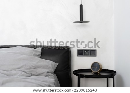 detail in bedroom interior, pillow and duvet in cotton case on bed with black leather headboard, alarm clock on metal coffee table and electrical socket with switch on white wall
