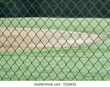 Detail of chain link fence in front of distant baseball field. Lush green grass and the clay or dirt of the diamond are visible. Beyond, at the edge of the field, is a dense cluster of green trees