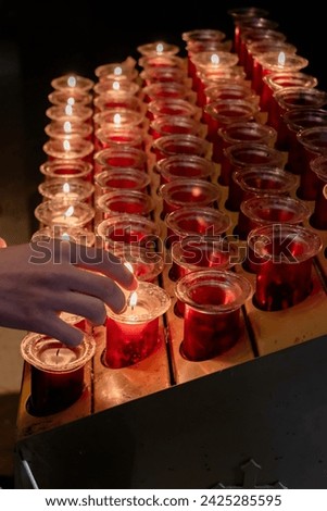 Detail of a child's hand lighting small devotional candles in small jars placed on a lectern in a Catholic cathedral. Small lighted candles represent the petitions of the parishioners. Vertical.
