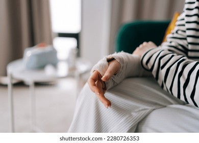 Detail cropped shot of suffering from pain unrecognizable young woman with broken hand wrapped in plaster bandage, gently massaging injured arm, sitting at home. Concept of insurance and healthcare.