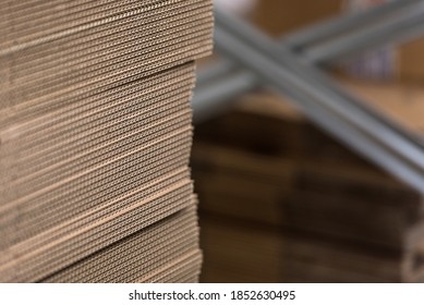 Detail Of Edge Of Neatly Stacked Corrugated Cardboard For Making Boxes, With Metal Factory Shelves In Background In Soft-focus