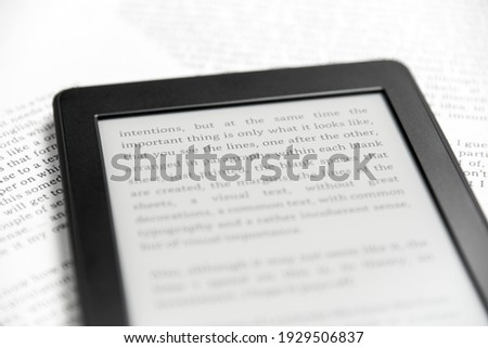 Detail of an electronic reader with the screen on and a text on it, on sheets of paper with text printed on them. Concepts: reading and digital technology.