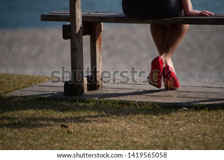 Detail of the feet of a girl sitting behind on a wooden bench. The girl is wearing a black skirt and red shoes with high heels. 