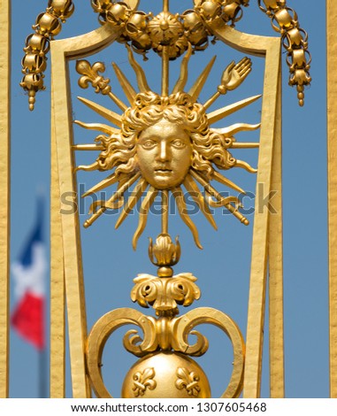 Detail of the gilded gates of the palace of Versailles outside Paris, showing the emblem of the Sun King, with a blue sky and the tricolour flag in the background.