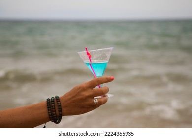 Detail Of Hand Raising Glass In The Air With A Blue Drink. In The Background You Can See The Sky And The Sea In The Horizon.