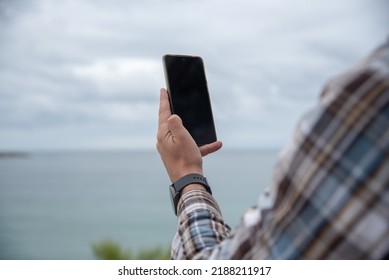 Detail of the hands of a young man taking a one-handed photograph of the landscape with his mobile phone in vertical on a cloudy day. Tourism, new technologies, development and lifestyle