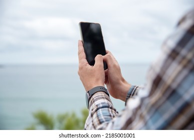 Detail Of The Hands Of A Young Man Wearing A Smart Watch On His Wrist And Typing On His Mobile Phone With A Landscape In The Background On A Cloudy Day.