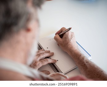 Detail of the head and the hands of an unrecognizable senior man writing in notebook at his desk - Powered by Shutterstock