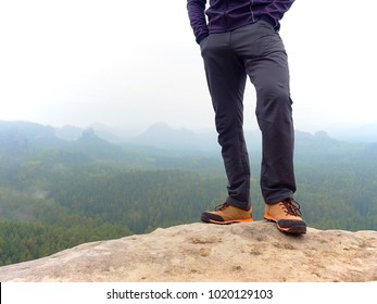 Detail Of  Hiker Legs In Black Orange Hiking Boots On Mountain Summit. Feets In Trekking Shoes And Legs  Light Trousers  On Background Of The Peaks