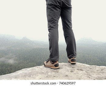 Detail Of  Hiker Legs In Black Orange Hiking Boots On Mountain Summit. Feet In Trekking Shoes And Legs  Light Trousers  On Background Of The Peaks