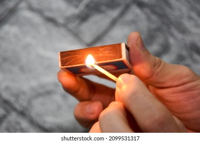 Detail Of Ignition Of Matches For A Matchbox. A Burning Match In One Hand And A Matchbox In The Other. The Process Of Lighting A Match. Hand With Burning Match On Gray Background