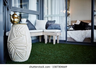 Detail Of An Inner City Apartment Balcony With Settees, Cushions And A Cup Of Tea And Tea Pot On A Side Table In Foreground, Bedroom Visible In Background