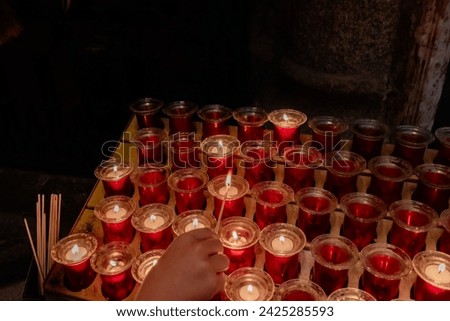 Detail of a kd's hand with a match lighting small devotional candles in small jars placed on a lectern in a Catholic cathedral. Small lighted candles represent the petitions of the parishioners.