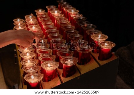 Detail of a kid's hand with a lighter lighting small devotional candles in small jars placed on a lectern in a Catholic cathedral. Small lighted candles represent the petitions of the parishioners.