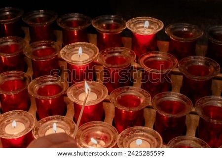 Detail of a kid's hand with a match lighting small devotional candles in small jars placed on a lectern in a Catholic cathedral. Small lighted candles represent the petitions of the parishioners.