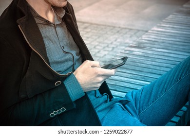 Detail of man with smartphone. He wears a blue jeans, a casual shirt and a black coat