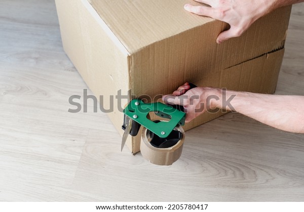 Detail of a man\
using a tool to seal a cardboard box with brown duct tape. Tape\
dispenser. Packing of moving\
boxes.