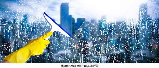 Detail on washer hand in yellow glove hold cleaning squeegee and clean dirty office window in huge skyscraper height. View on modern city as background. Cleaning service concept banner or wide