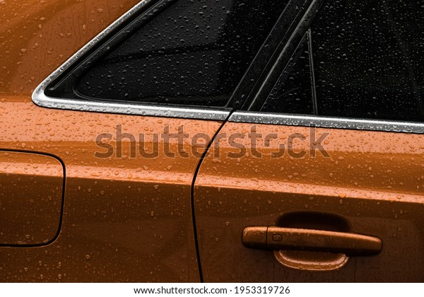 Detail of an\
orange car on a rainy day. Side window, door handle and orange car\
paint covered with\
raindrops.