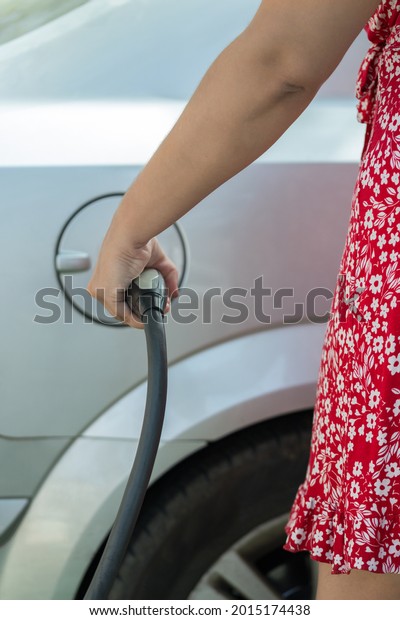 Detail\
of the power cable and plug of a charging station held by the left\
hand of a young woman seen from behind. She is approaching her\
electric car to start charging it. Vertical\
image.