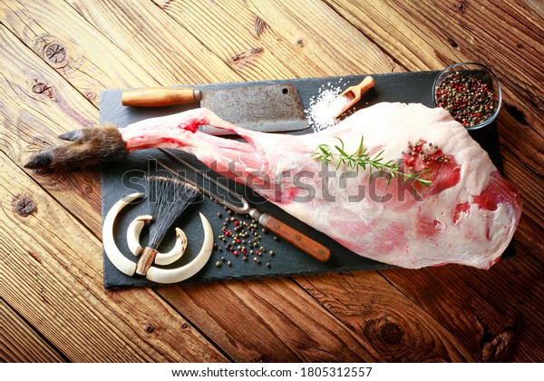 Detail of raw meat, thigh of wild boar on
a black plate with rosemary, salt and pepper. the atmosphere of
hunting is completed by weapons from wild
boar