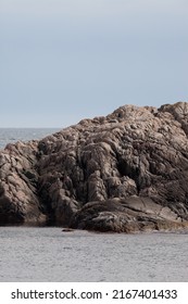 A detail of a rocky cliff surrounded by the sea. Rough rocks on the shore, sky and horizon in the background.