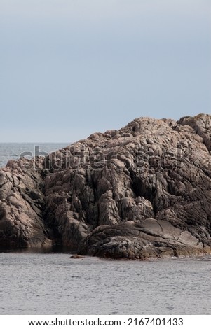 A detail of a rocky cliff surrounded by the sea. Rough rocks on the shore, sky and horizon in the background.