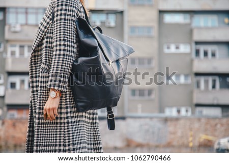 detail shot. attractive brunette woman outdoors posing in a checked patterned houndstooth coat, wearing a golden ring and watch and holding a black leather backpack. fashion outfit, streetstyle
