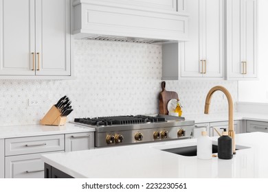 A detail shot of a beautiful kitchen's stainless steel luxury stove, hood, granite counter tops, and a custom, white and gold tiled back splash and faucet.