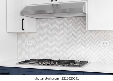 A Detail Shot Of A White And Blue Kitchen's Stainless Steel Stove Top, Granite Countertop, And A Marble Herringbone Tiled Backsplash.