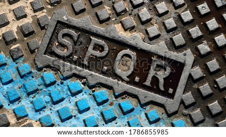 Detail of SPQR writing on the geometric cast iron manhole cover, with a strip of blue paint on a street in Rome