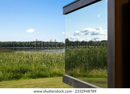 Detail view of modern mirror reflective window on building in the middle of wild nature. Luxury cottage concept. Lake, reed, trees, blue sky. 