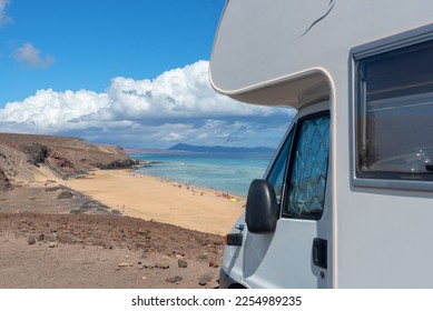 Detail of a white caravan, motorhome on a cliff of volcanic earth in front of a white sandy beach of turquoise sea during a sunny day on the coast of the touristic Fuerteventura in the Canary Islands.