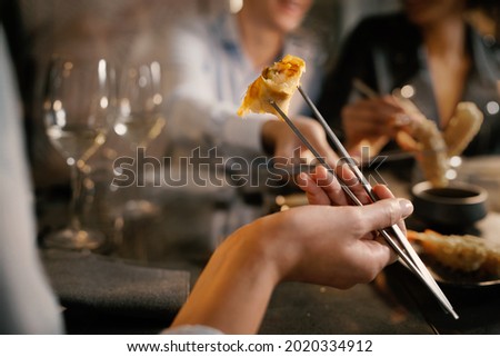 Detail of a woman's hand grabbing a spring roll with metallic chopsticks. He sits at a table with friends in a fancy asiatic fusion food restaurant.