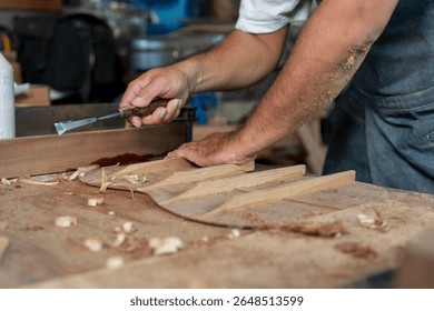 Detail of a woodworker shaping the interior bracing of a guitar with a chisel on a workbench. Sawdust covers arms and table. Concept of luthier craftsmanship, handmade process, and attention to detail - Powered by Shutterstock - Get 15% off with code: PIKWIZARD15