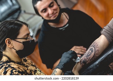 Detail Of Young Tattoo Artist Girl With Glasses And Mask Cleaning A New Tattoo Of 'La Santa Muerte' (Our Lady Of Holy Death) In The Arm Of A Woman And Apprentice Watching