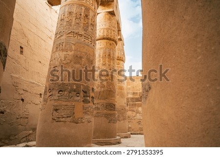 A detailed angled view of multiple columns in the Great Hypostyle Hall at Karnak Temple in Luxor, Egypt, showcasing vivid painted hieroglyphic reliefs with warm orange, red, and ochre tones. The carvings depict ritual scenes, deity figures, and royal cartouches preserved in remarkable condition after more than three millennia.