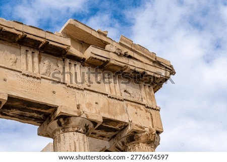 A detailed architectural shot of the upper section of the Parthenon temple, showing the Doric metopes and triglyphs of the frieze. The image highlights the ancient stone carvings and the weathering of the marble under a bright, cloudy sky in Greece.