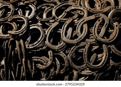 A detailed, close-up pattern of overlapping old metal horseshoes showing heavy rust and texture. The dark metallic pieces are piled closely together in natural sunlight.